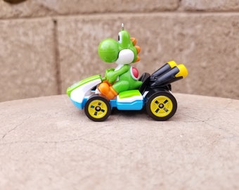 May include: A green Yoshi character driving a white, blue, and black race car with yellow wheels. The car has two black exhaust pipes.