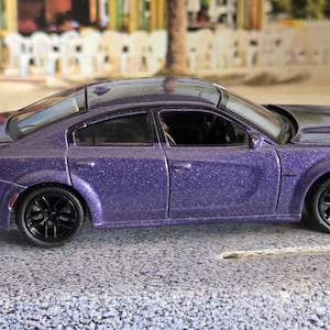 May include: A purple toy car with black wheels. The car has a sleek design with a spoiler and a hood scoop. The car is on a gray surface.