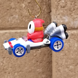 May include: A red and white toy car ornament with a red character with a white face and black eyes driving the car. The car has blue wheels and silver rocket boosters.