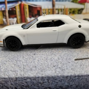 May include: A white toy car with black wheels and a black spoiler. The car has a sleek design and appears to be a model of a sports car. The car is set against a blurred background of a beach scene.