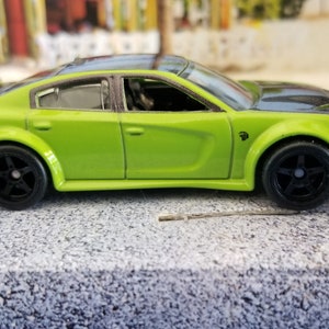 May include: A green toy car with black wheels and a black hood. The car has a sleek design and is sitting on a gray surface. The car is a Dodge Charger.