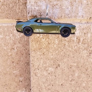May include: A metallic olive green toy muscle car ornament featuring a black rear spoiler, black wheels, and a detailed interior. The vehicle is suspended by a silver metal hook, showcasing a sleek coupe body style with defined side panels.