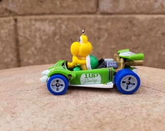 May include: A green and white toy race car with a yellow turtle character driving. The car has blue wheels and a gold exhaust pipe. The car has the words "1UP Racing" on the side.