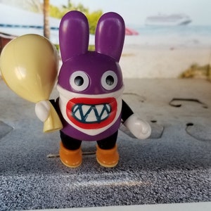 May include: A purple rabbit figurine with a white face, large teeth, and a yellow balloon. The figurine has orange boots and black pants. The background shows a beach scene.