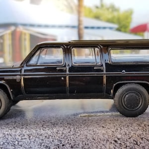 May include: Black toy model of a Chevrolet Suburban SUV. The model has a black body, black wheels, and a black interior. The model is in a side view.
