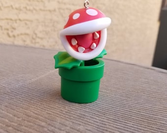 May include: A decorative ornament featuring a Piranha Plant from the Super Mario video game series. The plant has a red head with white spots, a white mouth with teeth, and is set in a green pot. The ornament includes a silver loop for hanging.