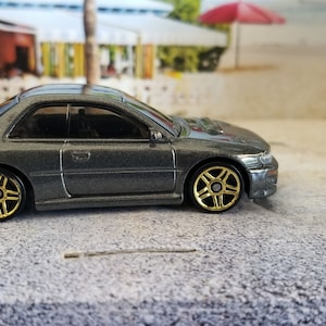 May include: A gray Subaru Impreza WRX STI diecast car model with gold wheels. The car is sitting on a gray, pebbled surface.