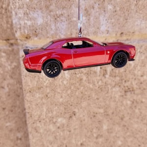 May include: A red toy car with black wheels and a spoiler. The miniature vehicle is suspended by a silver hook, showcasing its glossy finish and detailed design. The car is a coupe style.
