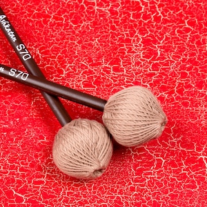 Może przedstawiać: Dwa brązowe drewniane młotki z beżowymi główkami z przędzy, oznaczone "Ashkatar S70", na czerwonej, popękanej powierzchni.