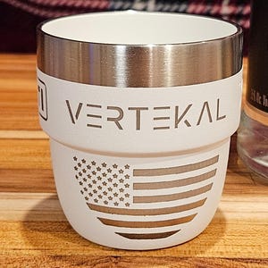 May include: White stainless steel travel mug with a silver band and the word "VERTEKAL" engraved on the side. The mug has an American flag design on the bottom.