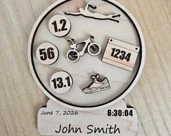 May include: A circular wooden ornament featuring laser-cut icons of a swimmer, a bicycle, and a running shoe. It includes circular discs with the numbers 1.2, 56, and 13.1, a rectangular bib with 1234, and a base engraved with June 7, 2026, 6:30:04, and John Smith.