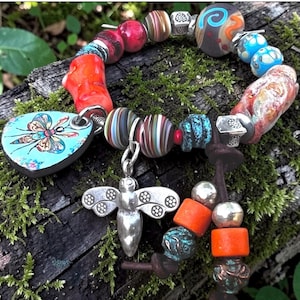 Op de afbeelding: Een handgemaakte armband met een verscheidenheid aan kralen in rood, blauw, oranje en zilver. De armband bevat een zilveren bijenbedel en een blauwe hartvormige hanger met een libellenontwerp. De armband wordt getoond op een mosachtig oppervlak.