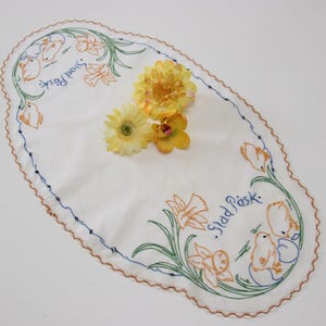 May include: Oval white embroidered Easter table runner with floral and chick designs. The border is scalloped with a brown trim and blue stitching. The text "Glad Påsk" is embroidered in blue. Yellow flowers are placed on top.