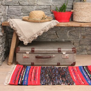 Può includere: Una valigia vintage con una maniglia in pelle marrone si trova su una panca di legno con una coperta all'uncinetto e un cappello di paglia. Un vaso di metallo rosso con una pianta verde si trova sulla panca. Un tappeto intrecciato con strisce rosse, blu e bianche è sul pavimento.