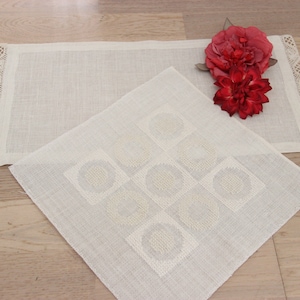 May include: Two white linen placemats with a delicate lace trim. The smaller placemat has a geometric pattern of circles. A pair of red silk flowers are placed on the larger placemat.