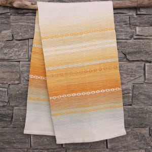 Puede incluir: Un paño de cocina plegado y tejido con un degradado de colores. La toalla pasa del blanco al amarillo, al naranja y a un toque de azul. Presenta rayas horizontales con diseños de diamantes y puntos. La toalla está colgada contra una pared de piedra gris.