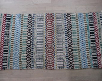 May include: A rectangular woven rug with a striped pattern. The rug features vertical stripes in various colors, including red, blue, black, and white. The rug has a fringed edge on both ends. The rug is made of a soft, textured material.