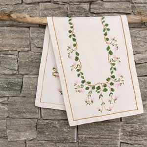 Könnte beinhalten: Ein weißer Tischläufer mit einem floralen Design mit rosa Blüten und grünen Blättern. Der Läufer hängt an einem Holzast an einer Steinwand.