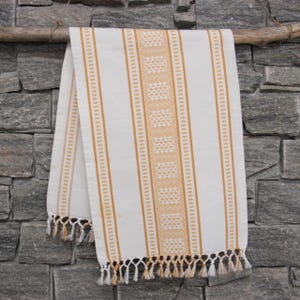 Puede incluir: Toalla decorativa a rayas blancas y doradas con una banda central estampada. La toalla cuelga de una rama de madera contra una pared de piedra gris. El borde inferior presenta una borla de flecos blancos y dorados.