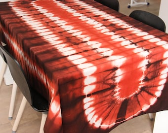 May include: Red, white, and black tie-dye tablecloth with a circular pattern in the center. The tablecloth is draped over a table with two black chairs.