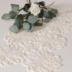 Puede incluir: Un camino de mesa de encaje blanco tejido a crochet con un borde festoneado y una rama de eucalipto con una rosa blanca.