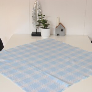Peut inclure: Une nappe carrée à carreaux bleu clair et blancs est posée sur une table blanche. La nappe a un bord effiloché. Des objets décoratifs, dont une plante, une bougie et un ornement en forme de maison, sont visibles en arrière-plan.