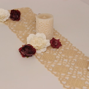 May include: A beige crocheted table runner with white and burgundy flowers and a white candle in the centre.