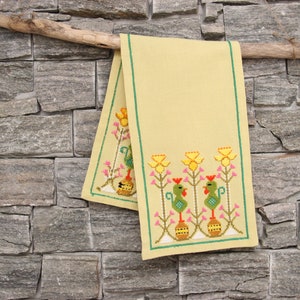 May include: A yellow linen table runner with embroidered green roosters and yellow flowers. The runner is hanging on a wooden branch against a stone wall.