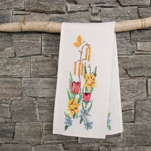 May include: A white linen tea towel with an embroidered floral design featuring yellow daffodils, red tulips, and blue forget-me-nots. The towel is hanging on a rustic wooden branch.