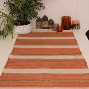 Puede incluir: Una mesa blanca con un camino de mesa a rayas en tonos naranja y marrón. El camino de mesa está hecho de un material tejido y tiene un aspecto rústico. Hay dos sillas negras a cada lado de la mesa y una planta en maceta en el fondo.