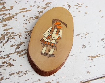 May include: A wooden oval-shaped plaque with a painted image of a person in a traditional costume holding a scythe. The person is wearing a white shirt with orange and black geometric patterns, orange pants, and a red hat. The background is a light brown wood.