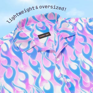 May include: A button-down shirt with a vibrant flame pattern in shades of blue, pink, and white. The shirt has a classic collar and is labeled "lightweight & oversized!" in white text. The shirt has a small label on the inside collar.