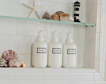 May include: White tiled bathroom shelf with three white pump bottles labeled "Shampoo", "Conditioner", and "Body Wash". The shelf also has a small bird figurine, seashells, and a starfish.