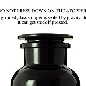 May include: A black glass bottle with a black glass stopper. The bottle has a round body and a narrow neck. The stopper is sealed by gravity alone. The text on the bottle reads: "DO NOT PRESS DOWN ON THE STOPPER! The grinded glass stopper is sealed by gravity alone. It can get stuck if pressed."