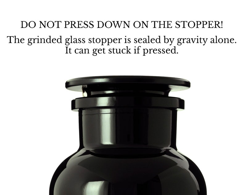 May include: A black glass bottle with a black glass stopper. The bottle has a round body and a narrow neck. The stopper is sealed by gravity alone. The text on the bottle reads: "DO NOT PRESS DOWN ON THE STOPPER! The grinded glass stopper is sealed by gravity alone. It can get stuck if pressed."