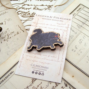 May include: A wooden badge pin featuring a black and red illustration of a mythical creature. The pin is labelled "Legend & Folklore Wooden Badge Pin" and includes the artist's name and website.