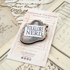 May include: Wooden badge pin with a folklore theme. The pin features a mythical creature holding a sign that reads "FOLKLORE NERD." The pin is on a card with the text "LEGEND & FOLKLORE - WOODEN BADGE PIN."