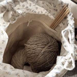 Puede incluir: Una bolsa con cord&oacute;n de color crema con un estampado floral azul, abierta para revelar dos ovillos de hilo marr&oacute;n y agujas de tejer de madera. El interior de la bolsa es de color beige claro y se ve un bolsillo con cremallera.