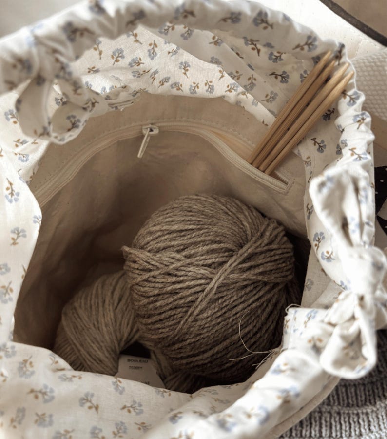 Puede incluir: Una bolsa con cord&oacute;n de color crema con un estampado floral azul, abierta para revelar dos ovillos de hilo marr&oacute;n y agujas de tejer de madera. El interior de la bolsa es de color beige claro y se ve un bolsillo con cremallera.