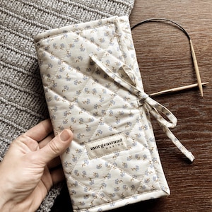 Puede incluir: Un estuche acolchado para agujas de tejer de color crema, con un estampado floral y una cinta a juego. El estuche tiene una etiqueta que dice "morgenstund making". Tambi&eacute;n se ven agujas de tejer de madera y un art&iacute;culo de punto gris.