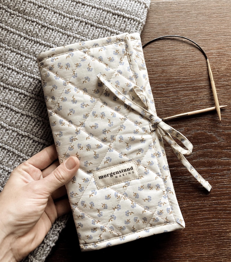 Puede incluir: Un estuche acolchado para agujas de tejer de color crema, con un estampado floral y una cinta a juego. El estuche tiene una etiqueta que dice "morgenstund making". Tambi&eacute;n se ven agujas de tejer de madera y un art&iacute;culo de punto gris.