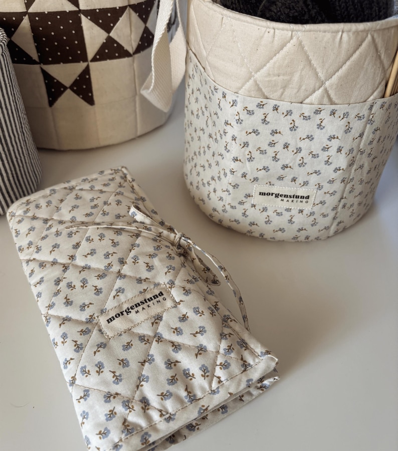 Puede incluir: Una colecci&oacute;n de cestas de almacenamiento y un estuche de tela acolchada. Las cestas y el estuche son de color crema con un estampado floral azul. El estuche est&aacute; atado con una cinta color crema. El texto "morgenstund making" es visible.