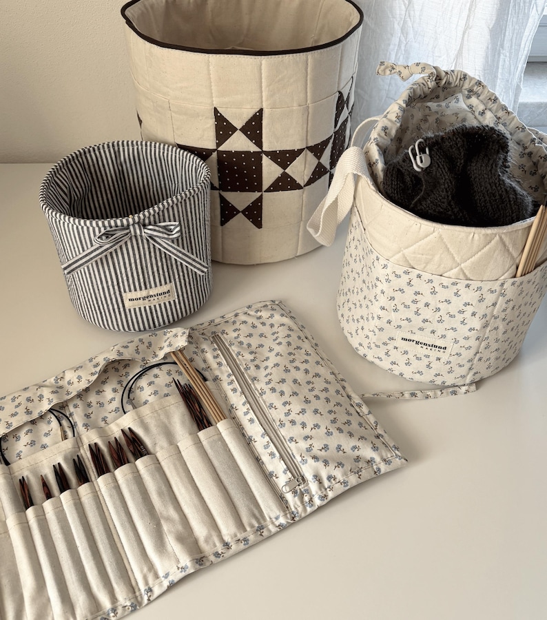 Puede incluir: Una colecci&oacute;n de contenedores de almacenamiento de tela hechos a mano y un estuche para agujas de tejer. Los contenedores incluyen una cesta a rayas con un lazo, una bolsa con cord&oacute;n con estampado floral y una cesta m&aacute;s grande con un dise&ntilde;o de estrella. El estuche de agujas est&aacute; abierto, mostrando varias agujas de tejer.
