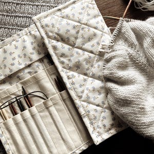 Puede incluir: Un estuche acolchado de color crema para agujas de tejer con un estampado floral, abierto para revelar varias agujas de tejer. Se ve un su&eacute;ter gris parcialmente tejido y lana, lo que sugiere un proyecto de tejido. El estuche est&aacute; hecho de tela y tiene varios bolsillos.