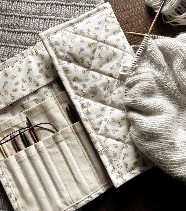 Puede incluir: Un estuche acolchado de color crema para agujas de tejer con un estampado floral, abierto para revelar varias agujas de tejer. Se ve un su&eacute;ter gris parcialmente tejido y lana, lo que sugiere un proyecto de tejido. El estuche est&aacute; hecho de tela y tiene varios bolsillos.