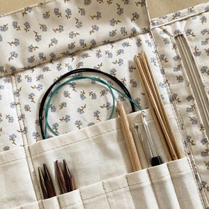 Puede incluir: Un estuche de agujas de tejer color crema con un estampado floral, que contiene varias herramientas de tejido. El estuche est&aacute; abierto, revelando agujas de tejer de madera, ganchillos y agujas circulares. La tela tiene un dise&ntilde;o floral azul.