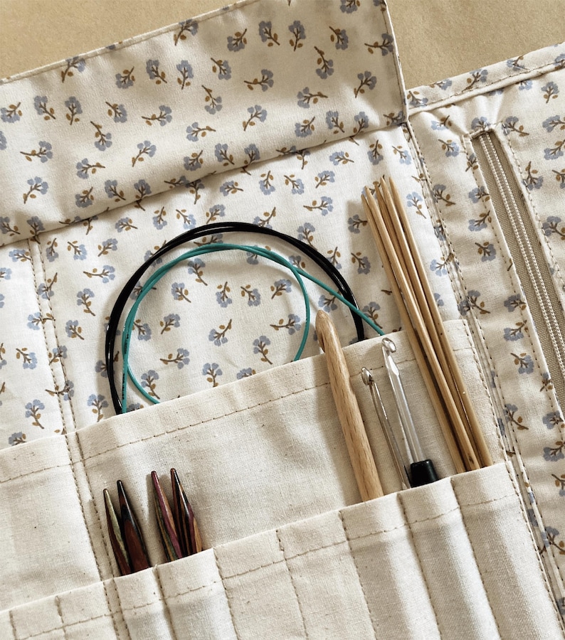 Puede incluir: Un estuche de agujas de tejer color crema con un estampado floral, que contiene varias herramientas de tejido. El estuche est&aacute; abierto, revelando agujas de tejer de madera, ganchillos y agujas circulares. La tela tiene un dise&ntilde;o floral azul.