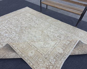 May include: A large, rectangular beige rug with an intricate, faded floral pattern. The rug has a detailed border in a darker beige color. The rug is partially rolled up, revealing its underside. The rug is placed on a dark surface.