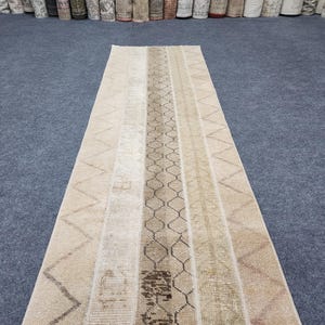 Peut inclure: Un long tapis de couloir rectangulaire présentant un motif patchwork de bandes verticales dans des tons beige, crème et brun clair. Le design comprend des sections avec des lignes en zigzag et une bande centrale à motif hexagonal.