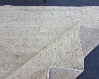 Può includere: Un tappeto beige e crema con un intricato motivo floreale e un bordo decorativo. Il tappeto è parzialmente piegato, rivelando il lato inferiore, ed è posto su una superficie blu scuro.